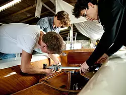 Drei Schüler arbeiten im Bootsschuppen am Traveller einer Holzjolle.